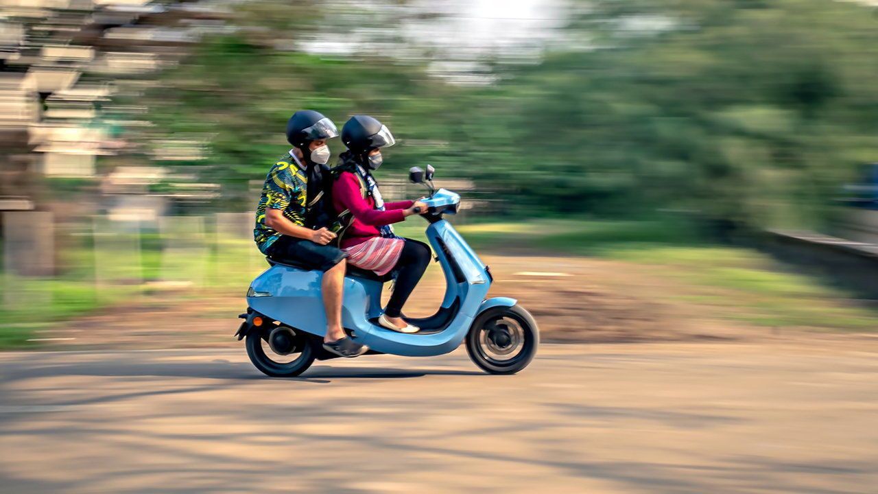 A couple wearing helmets riding on a blue Ola electric moped.